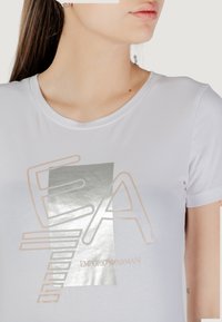 Camiseta de algodón blanca con un diseño gráfico metálico en plata y beige que presenta el texto "EA" y "Emporio Armani" en el centro. Mangas cortas.