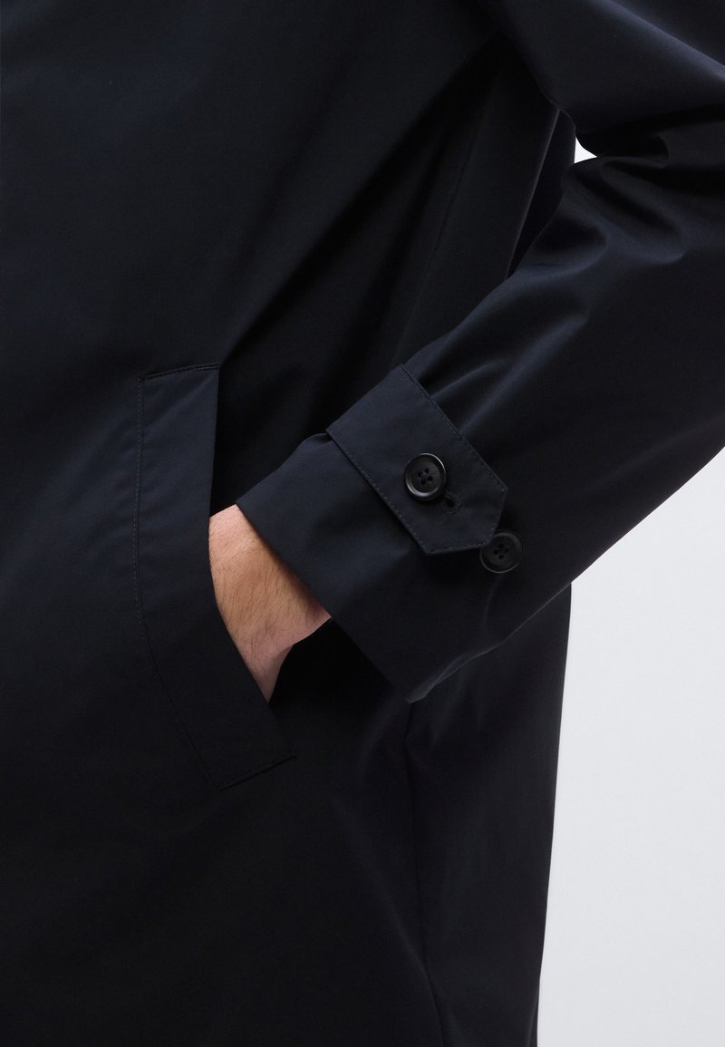 Black jacket with a smooth, matte finish. Features a chest pocket and buttoned cuffs. Cuffs have dual buttons and a clean design.