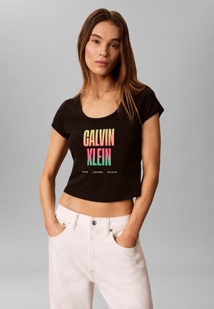 Femme aux longs cheveux bruns porte un t-shirt court noir avec un logo coloré Calvin Klein et un pantalon blanc, mains dans les poches, fond neutre.