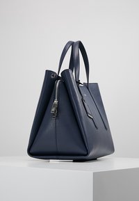 Granatowa skórzana torba na ramię z podwójnymi uchwytami, srebrnym zamkiem błyskawicznym i strukturalnym trójkątnym wzorem po bokach na białej powierzchni.