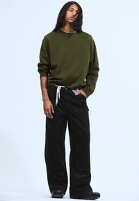 Person mit langen dunklen Haaren, die einen olivgrünen Sweatshirt, schwarze weite Hosen und schwarze Schuhe trägt, steht vor einem schlichten hellen Hintergrund.
