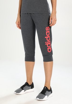 Onderbenen van een persoon die donkergrijze Adidas capri broek draagt met een rood logo op het rechterbeen en zwart-grijze slip-on sneakers met witte zolen.