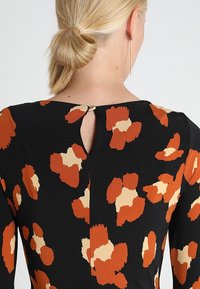 Robe noire avec un motif floral abstrait orange et beige. Présente un détail de dos en découpe en cœur et une texture lisse et douce.