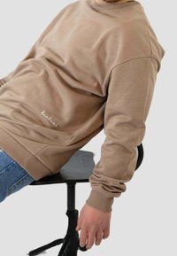 Persona con un sudadera beige que lleva el texto "luckluck", sentada de lado en una silla de oficina negra, con un brazo relajado hacia abajo.