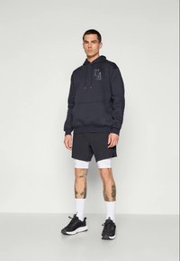 Mand står iført en sort hættetrøje med "UA" logo, sorte shorts over hvide kompressionsleggings, hvide sokker og sorte sneakers foran en ensfarvet baggrund.