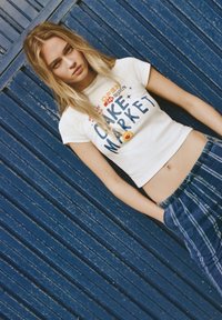 Hvid kortærmet t-shirt med teksten og grafikken "Cake Market". Sammen med blå ternede bukser. Blå træbaggrund.