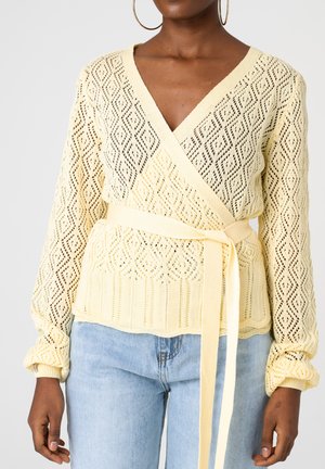 Frau trägt einen gelben Strick-Wickelpullover mit Rautenmuster, der an der Taille gebunden ist, kombiniert mit hellblauen Jeans und großen Creolen.