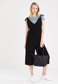 Schwarzer Jumpsuit mit Schleifenakzenten an den Trägern, über einem hellblauen T-Shirt getragen. Kombiniert mit einer schwarzen Tragetasche und weißen Sneakers.