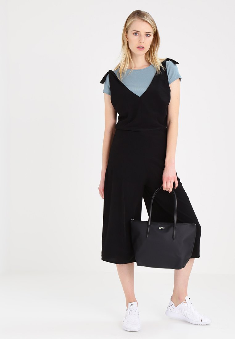 Schwarzer Jumpsuit mit Schleifenakzenten an den Trägern, über einem hellblauen T-Shirt getragen. Kombiniert mit einer schwarzen Tragetasche und weißen Sneakers.