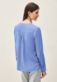 Camicia azzurra a maniche lunghe realizzata in tessuto morbido, con pince sul retro, orlo arrotondato e polsini con bottone.