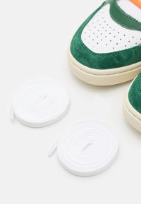 Zielone zamszowe trampki z białymi skórzanymi akcentami i perforacjami. Obok butów leżą dwie okrągłe białe sznurówki.
