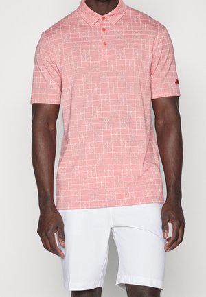 Uomo che indossa una polo rosa a maniche corte con motivo a griglia geometrica bianca e pantaloncini bianchi, in piedi contro uno sfondo semplice.