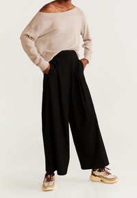 Beiger, schulterfreier Strickpullover, lässige schwarze weite Hose und klobige Schuhe mit beige-silbernen Akzenten. Glatte Texturen überall.