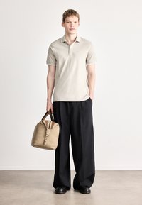 Μπεζ πόλο πουκάμισο με λευκό γιακά, φορεμένο με άνετο μαύρο παντελόνι. Το μοντέλο κρατά μια καφέ τσάντα με φερμουάρ και καφέ ρίγες. Μαύρα παπούτσια.