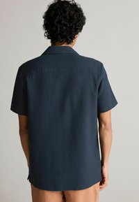 Camisa de manga corta en azul marino oscuro con tela texturizada, cuello puntiagudo y un patrón sutil. Bajo rematado con aberturas laterales.
