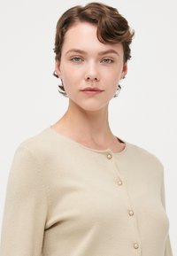 Beige strikket cardigan med rund hals og fem perle-lignende knapper på fronten. Glatt tekstur med en tettsittende design.