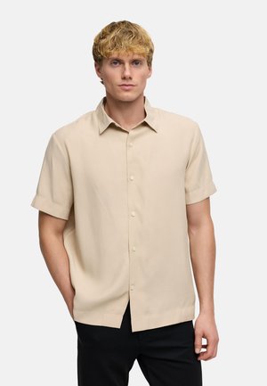 Ung mand med blondt hår iført en kortærmet beige skjorte med knapper og sorte bukser, stående med den ene hånd i lommen.