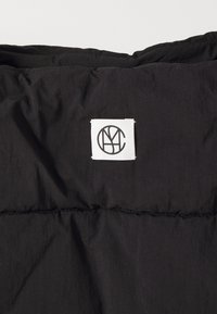 Svart vadderad tyg med en sydd design, med en fyrkantig vit etikett med en svart logotyp i det övre hörnet.