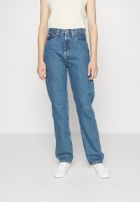 High-waisted blue denim jeans with a relaxed fit, featuring five pockets and a zip-fly closure, worn with white sneakers.