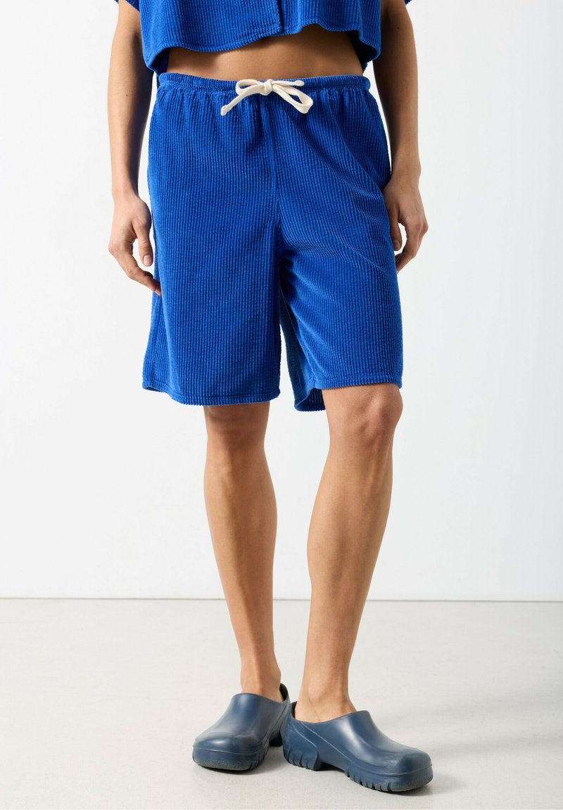 Niebieskie sztruksowe szorty z elastycznym pasem i sznurkiem do ściągania, o miękkiej fakturze. Do tego granatowe wsuwane chodaki.