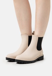 Botas de lluvia beige hasta el tobillo con paneles elásticos laterales negros y suelas gruesas de goma negra, usadas sobre piernas desnudas.