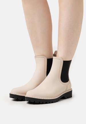 Beige kotníkové gumové boty na dešť se černými elastickými bočními panely a silnými černými gumovými podrážkami, nošené na holých nohách.