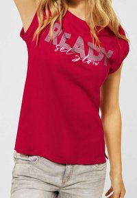 Röd t-shirt med kort ärm, gjord av mjukt tyg, med texten "READY set glow" i vitt och grå ränder, med en avslappnad passform och rundad fåll.