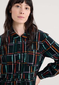 Kombinezon s dugim rukavima u crnoj tkanini s raznobojnim geometrijskim uzorkom u zelenoj, crvenoj, bijeloj i ljubičastoj boji; uključuje dugmad i ovratnik.