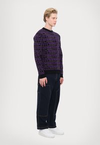 Purple knit sweater with black trim featuring a repeated text pattern, paired with navy cargo pants and white sneakers.