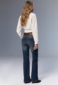 Vit tröja och blå utställda jeans med ett brunt läderbälte. Håret är vågigt och långt. Bakgrunden är ljusgrå.