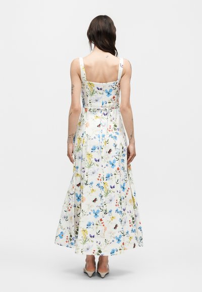 Γυναίκα που φοράει λευκό αμάνικο μακρύ φόρεμα με πολύχρωμο σχέδιο λουλουδιών και πεταλούδων, στέκεται με την πλάτη προς τα εμπρός σε απλό φόντο.