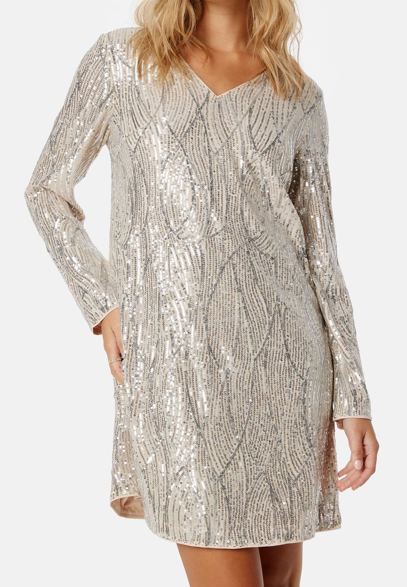 Femme portant une robe argentée à manches longues et col en V, couverte de motifs verticaux et ondulés en sequins, main reposant dans la poche.