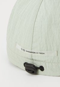 Chapeau vert clair en tissu texturé avec un motif en quadrillage, doté d'un cordon de serrage noir et d'une étiquette blanche portant la mention "CHAMPION C-TECH."