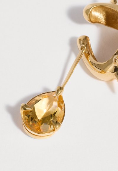 Orecchino in oro con una gemma gialla a forma di goccia sostenuta da tre griffes su un sottile gancio d'oro, su sfondo bianco.