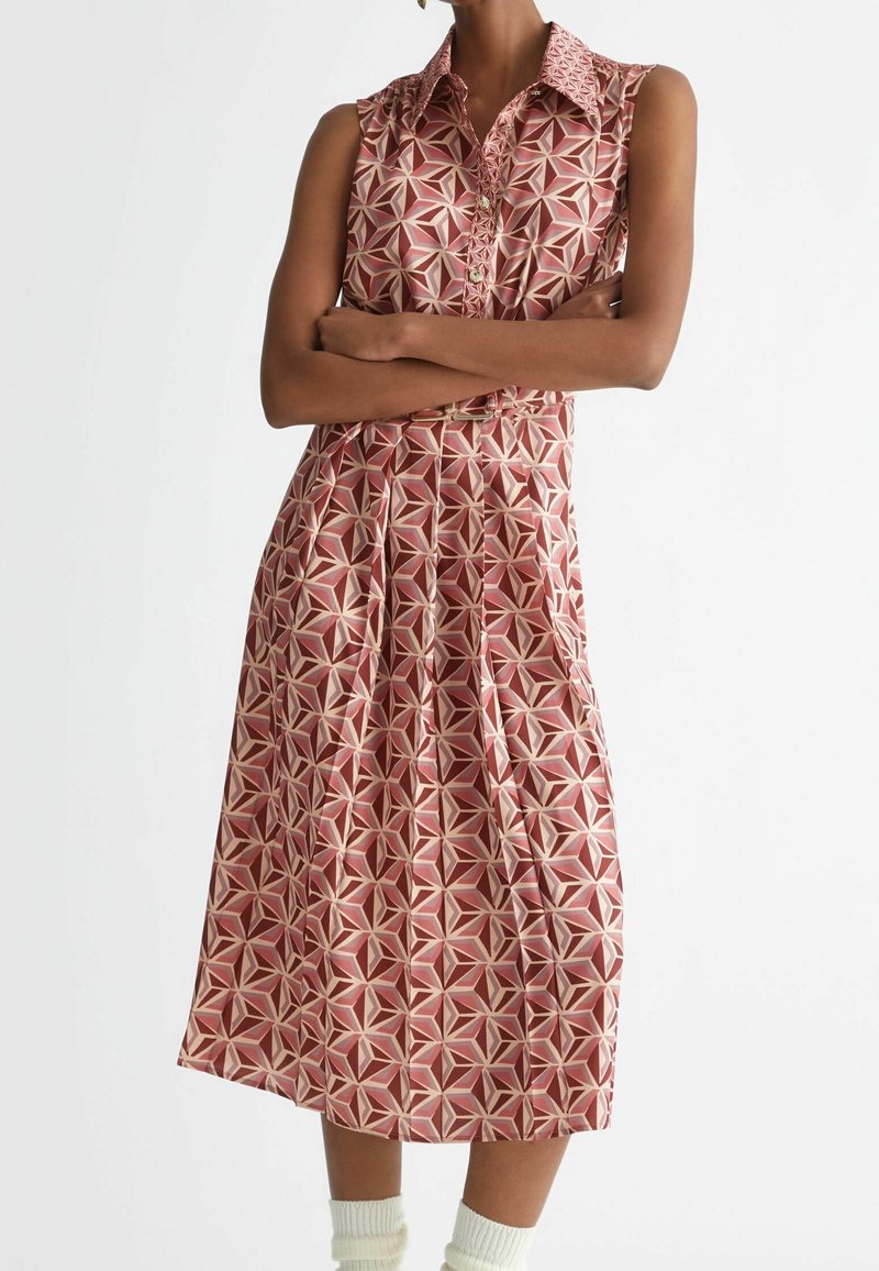 Femme debout, les bras croisés, portant une robe sans manches, à col, longueur genou, à motif géométrique rouge et beige.