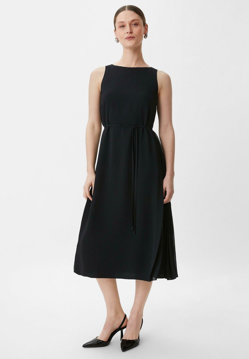 Schwarzes ärmelloses Kleid mit einem taillierten Oberteil, fließendem Rock und einem Band in der Taille. Kombiniert mit schwarzen spitz zulaufenden High Heels.
