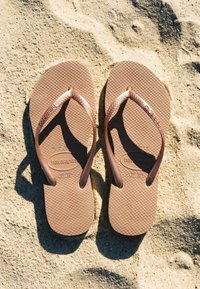 Καφέ λαστιχένια σαγιονάρες Havaianas, τοποθετημένες δίπλα-δίπλα σε ανοιχτόχρωμο αμμώδες έδαφος με υφή.