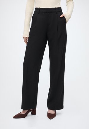 Femme portant un pantalon noir à jambes larges, un haut beige côtelé à manches longues et des chaussures marron pointues à petit talon, une main dans la poche.