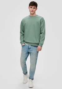 Felpa verde in cotone con maniche lunghe, scollatura tonda e polsini a costine, abbinata a jeans azzurri e sneakers bianche.