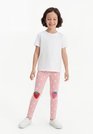 Bambino in piedi, indossa una maglietta bianca a maniche corte, leggings rosa con piccoli fiori bianchi e toppe a forma di fragola sorridente sulle ginocchia, scarpe bianche.