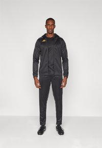Black athletic tracksuit made of smooth fabric. Zip-up hooded jacket with logo, paired with tapered pants and black sports shoes.