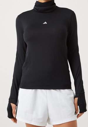 Woman wearing a black long-sleeve turtleneck top with thumb holes and white athletic shorts with Adidas logos.