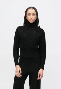 Crni pleteni džemper s visokim ovratnikom s teksturiranim uzorkom pletenice, rebrastim manžetnama i rubom. Uparen s crnim hlačama, nosi ga model.