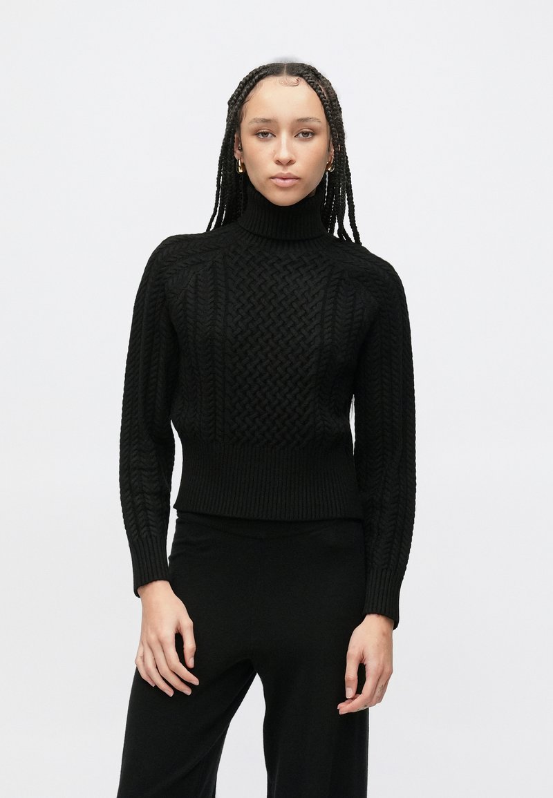 Crni pleteni džemper s visokim ovratnikom s teksturiranim uzorkom pletenice, rebrastim manžetnama i rubom. Uparen s crnim hlačama, nosi ga model.
