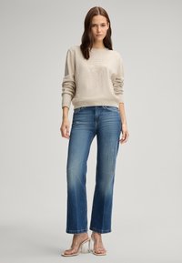 Maglione beige lavorato a maglia con un emblema testurizzato, abbinato a jeans flare chiari. Sandali con tacco trasparente completano l'outfit.