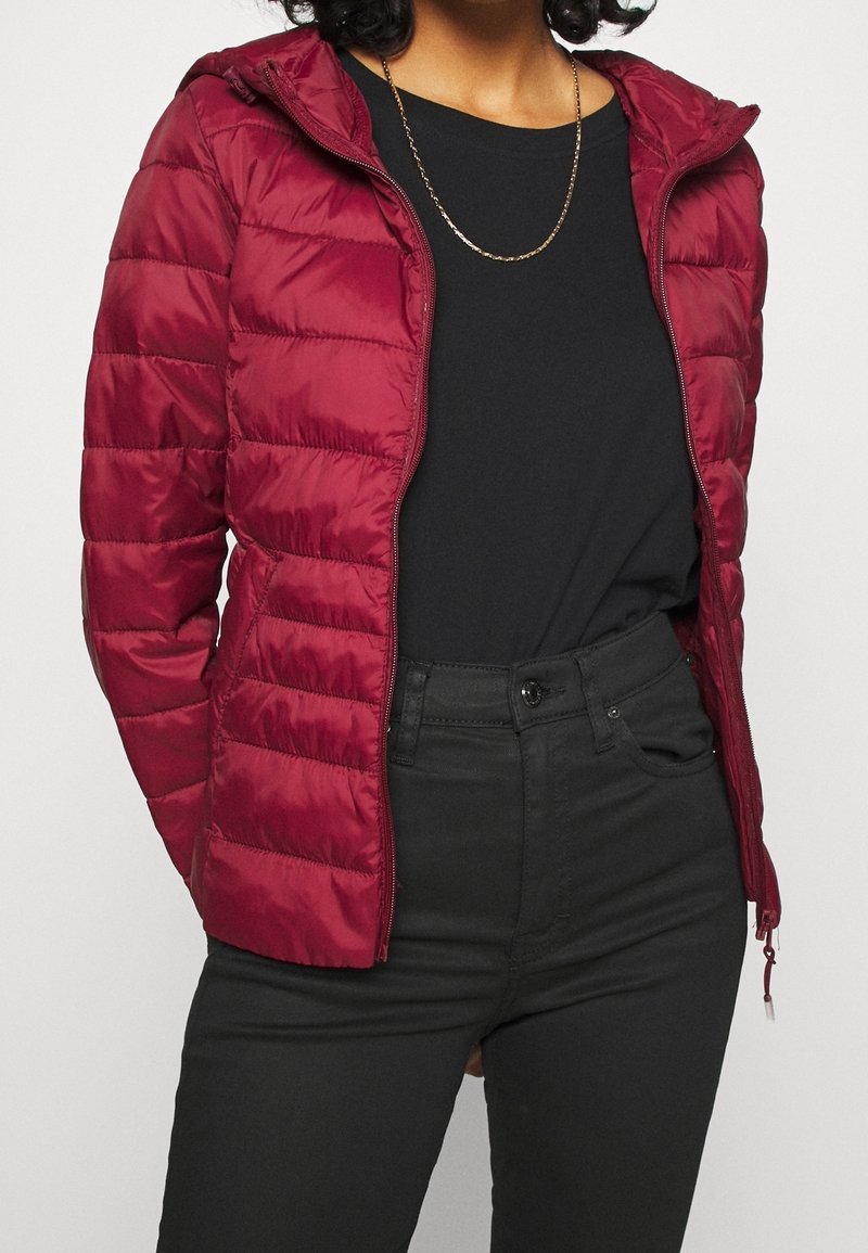 Rød dunjakke med hætte, med lynlås foran, quiltet mønster og glat tekstur. Båret over en sort top og tætsiddende sorte bukser.