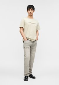 Ανοιχτό μπεζ μπλουζάκι με ανάγλυφο λογότυπο «Michael Kors», συνδυασμένο με ανοιχτό γκρι τζιν και μαύρα παπούτσια. Απλός σχεδιασμός, κανονική εφαρμογή και κοντά μανίκια.