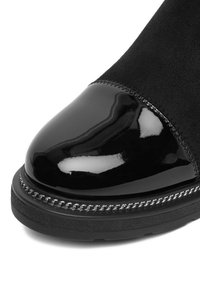 Botín negro de ante hasta el tobillo con una puntera de charol brillante y un acento de cadena plateada a lo largo de la suela. La parte superior texturizada contrasta con el acabado suave.