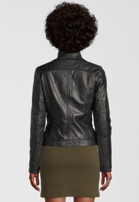 Zwarte leren jas met een strak ontwerp, hoge kraag en zichtbare stikdetails. Draagt een getextureerde olijfgroene jurk eronder.