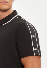 Camisa polo negra con un color blanco en el cuello y franjas laterales blancas en contraste, presentando un patrón de logotipos repetido en las mangas.